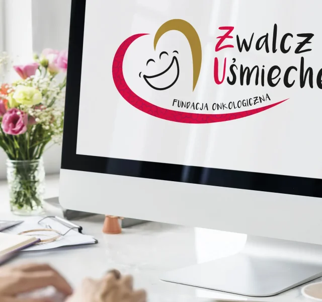 Logo Fundacji Zwalcz Go Uśmiechem wyświetlone na ekranie komputera w jasnym biurze, w tle kwiaty i notatnik na biurku. Fundacja wspierana przez KREOO - Studio projektowe z Mielca.