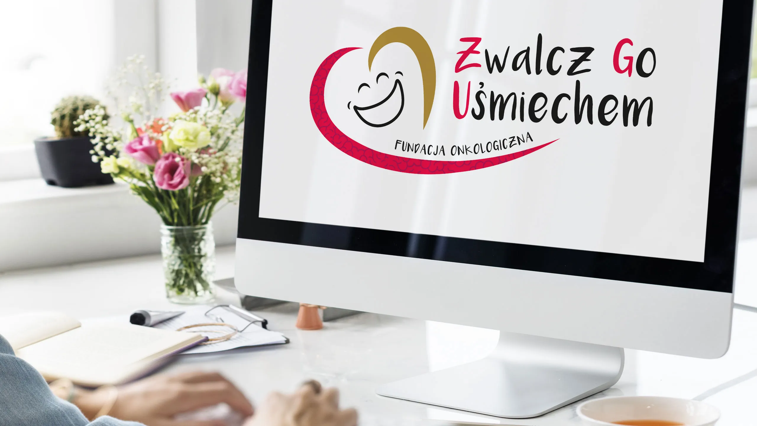 Logo Fundacji Zwalcz Go Uśmiechem wyświetlone na ekranie komputera w jasnym biurze, w tle kwiaty i notatnik na biurku. Fundacja wspierana przez KREOO - Studio projektowe z Mielca.