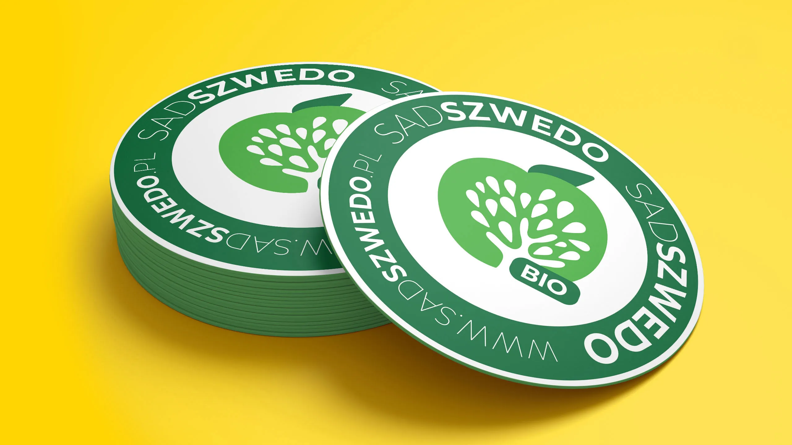 Okrągłe zielono-białe naklejki SadSzwedo BIO z symbolem jabłka i drzewa ułożone w stosie na żółtym tle
